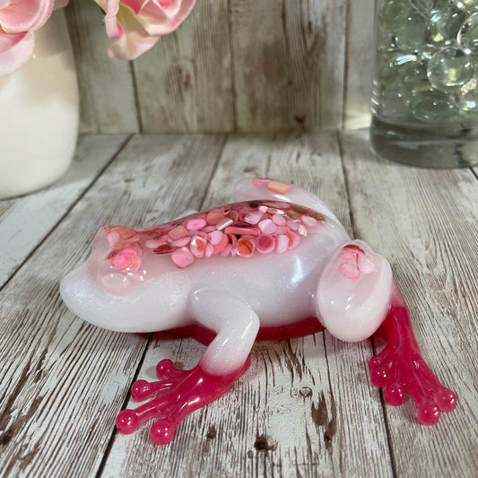 Decorative resin frog with pink accents on a wooden surface with flowers.